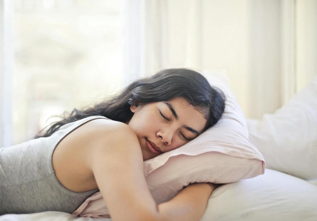 Woman laying in bed, her expression is serene and relaxed, she's hugging a pillow and she's wearing a grey tanktop. 